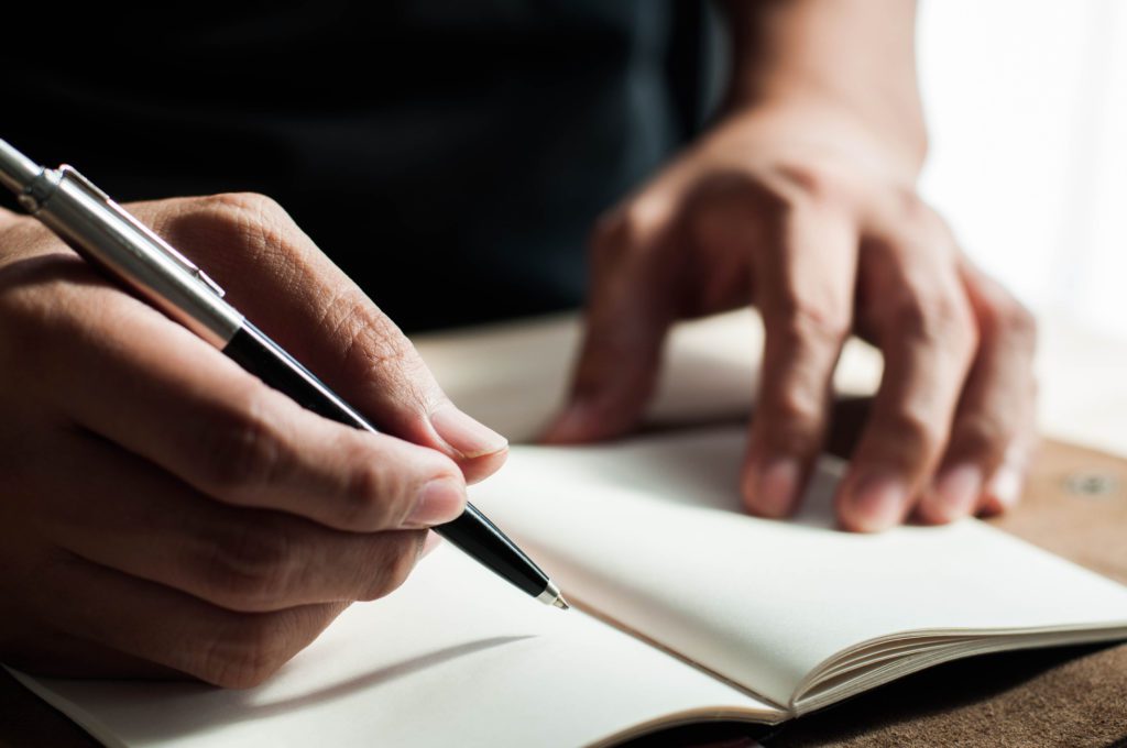 Image of a man's hand, holding a black pen, writing on a notebook.
